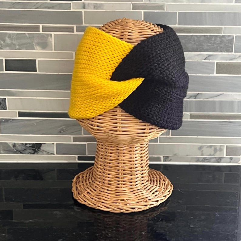 May include: A knitted headband featuring a two-tone design with one side in bright yellow and the other side in solid black. The fabric is constructed with a visible knit texture and includes a central twist detail where the two colours meet.