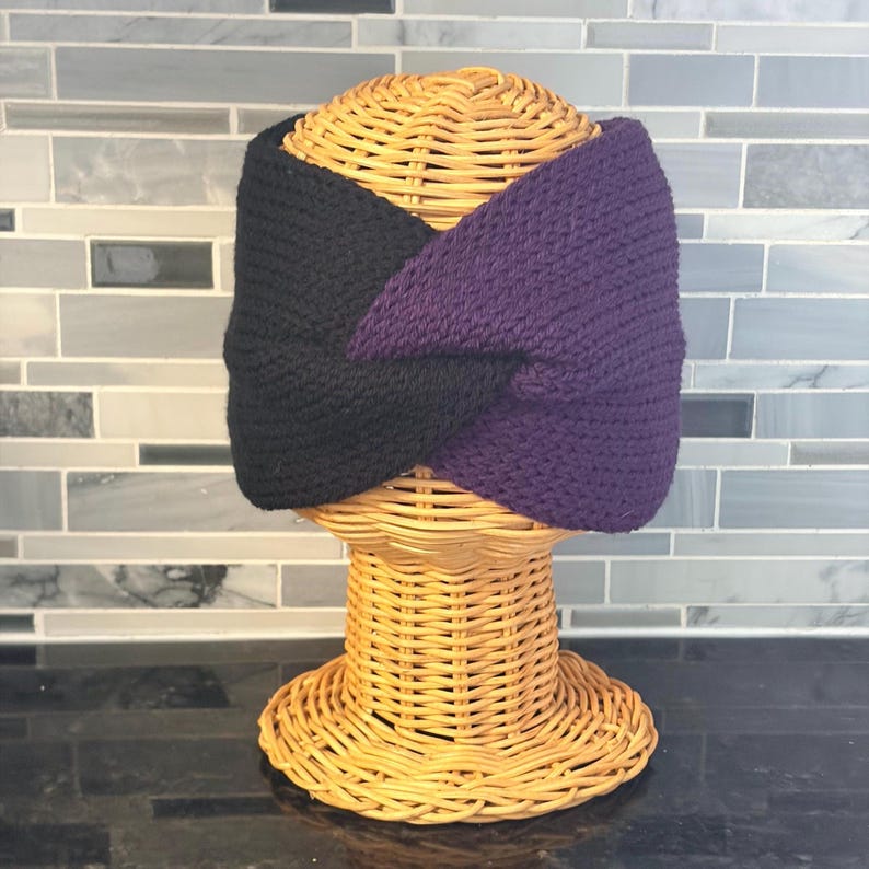 a black and purple knit headband displayed on a wicker head form. The headband is a twisted design, with one side black and the other a deep purple. The head form is made of woven wicker and sits on a matching base.