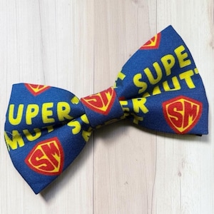 May include: A blue bow tie featuring a repeating pattern of yellow text that reads SUPER and red shield-shaped emblems containing the yellow letters SM. The fabric has a textured appearance and is arranged in a classic bow tie shape with two wings.