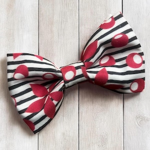 May include: A bow tie featuring a pattern of thin horizontal black stripes on a white background, accented with scattered red circular and petal-shaped motifs. The fabric has a visible woven texture, and the bow is tied in a classic shape.