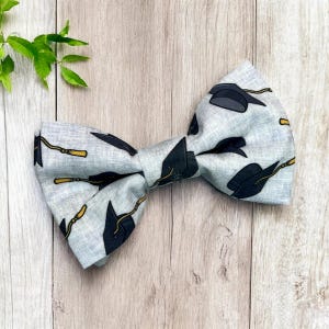 May include: A light grey bow tie featuring a pattern of black graduation caps and gold tassels. The bow tie is made of fabric and is tied in a classic bow shape. The background is a light brown wooden surface.