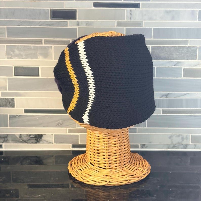 A black crochet hat with a curved design, featuring a yellow and white stripe. The hat is displayed on a wicker head form, showcasing its shape and texture. The hat is a stylish accessory.