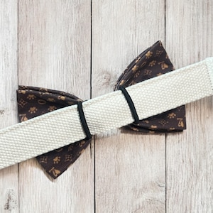 May include: A brown bow tie featuring a repeating gold pattern of paw prints and bone shapes. The bow tie is secured to a wide, off-white, textured fabric strap by two thin black elastic bands that hold the fabric in a gathered, folded shape.