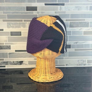 A knitted headband in purple, black, gold, and white, displayed on a wicker head form. The headband features a color-blocked design with a solid purple section and a black section with gold and white stripes.