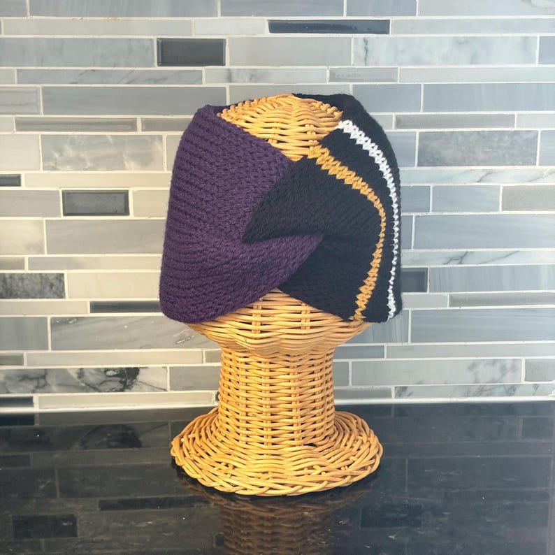 A knitted headband in purple, black, gold, and white, displayed on a wicker head form. The headband features a color-blocked design with a solid purple section and a black section with gold and white stripes.