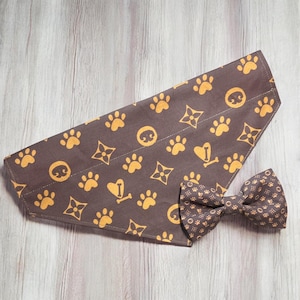 May include: A brown pet bandana and a matching bow tie featuring a repeating pattern of yellow paw prints, stylized four-pointed floral shapes, and heart-shaped icons with a small bone in the center, all printed on a dark brown fabric base.