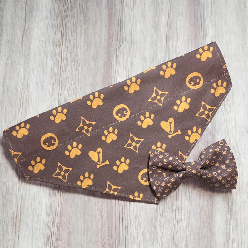 May include: A brown pet bandana and a matching bow tie featuring a repeating pattern of yellow paw prints, stylized four-pointed floral shapes, and heart-shaped icons with a small bone in the center, all printed on a dark brown fabric base.