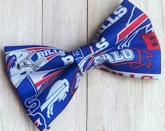 May include: A blue bow tie featuring a repeating pattern of white and red Buffalo Bills football team logos, including the charging buffalo silhouette, helmet graphics, and the word Bills in bold white lettering across the fabric surface.
