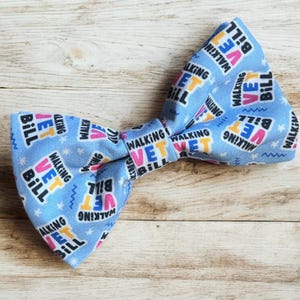 May include: A blue bow tie with a repeating pattern of "Walking Vet Bill" in pink, yellow, and black text. The bow tie is on a light brown wooden surface.