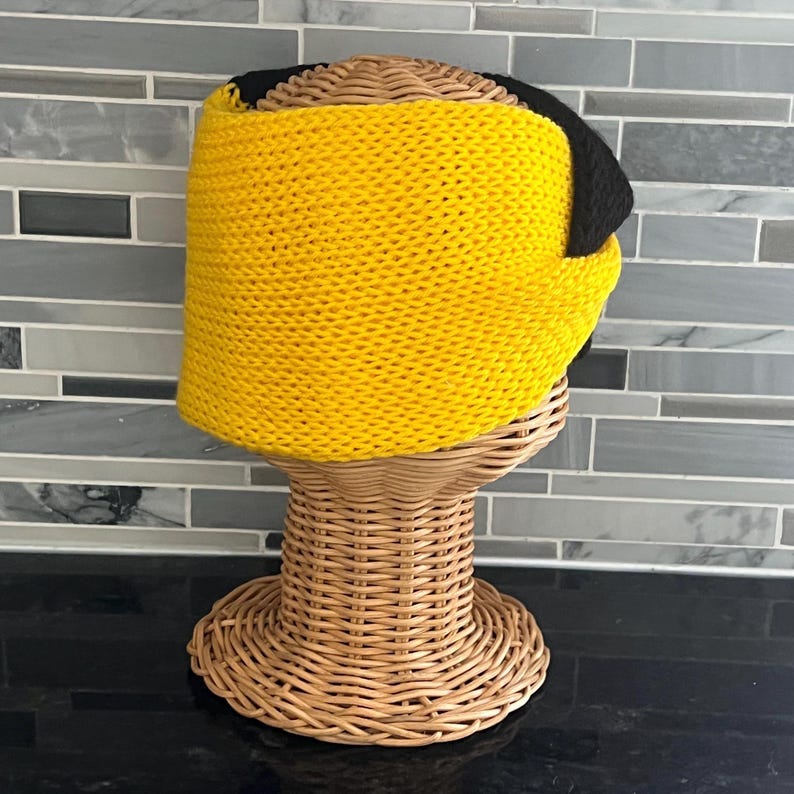 May include: A wide, knitted headband featuring a vibrant yellow front section and a contrasting black back section. The accessory is crafted with a textured, chunky knit pattern and is displayed on a woven wicker mannequin head stand.