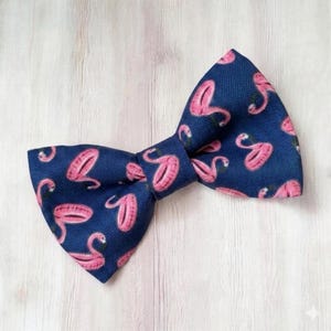 May include: A navy blue bow tie with a repeating pattern of pink flamingos. The bow tie is tied in a classic style and is made of a textured fabric. The background is a light wood grain.