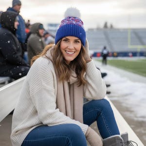 May include: A blue knit beanie with a white pom-pom and red and white patterned band, worn with a cream-colored ribbed sweater, a matching cream scarf, beige knit mittens, and blue jeans, while sitting in a snowy outdoor stadium.