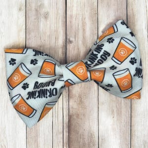 May include: A light gray bow tie featuring a repeating pattern of orange beer glasses with paw prints inside and the black text Drinking Buddy scattered across the fabric. The bow tie has a structured shape with a central knot and two wings.