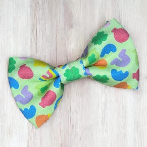 May include: A light green bow tie with a repeating pattern of colorful abstract shapes in pink, blue, orange, and green. The bow tie is centered on a light wood grain background.