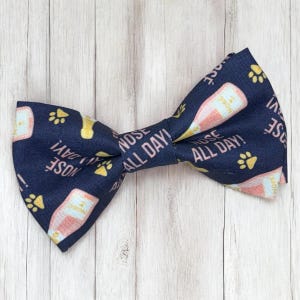 May include: A dark blue bow tie featuring a repeating pattern of light pink champagne bottles and yellow paw prints. The fabric is printed with the text "NOSE ALL DAY!" in a light pink font, arranged in a diagonal orientation across the surface.