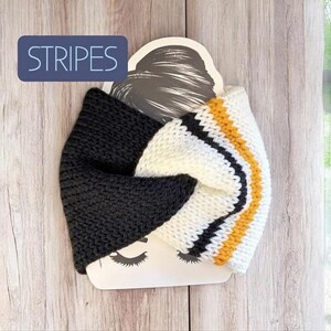 May include: A knitted headband featuring a twisted front design. One side is solid charcoal grey, while the other side displays white, black, and mustard yellow stripes. The headband is displayed on a card with the word STRIPES printed in white.