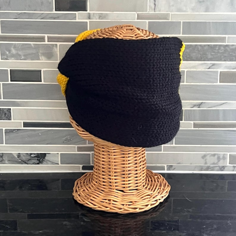 May include: A thick, hand-knitted headband featuring a wide black central band with vibrant yellow accents visible at the top and bottom edges. The textured, chunky yarn construction creates a dense, warm appearance on a woven wicker display stand.