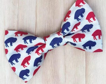 May include: A white bow tie featuring a repeating pattern of small, stylized buffalo silhouettes in alternating red and navy blue colors. The fabric has a crisp, clean appearance with the animal shapes distributed evenly across the entire surface.