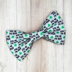 May include: A light blue bow tie featuring a repeating pattern of green shamrocks and the black text "KISS ME I'M IRISH". The fabric has a visible woven texture, and the bow tie is shaped with two distinct wings gathered at the center.