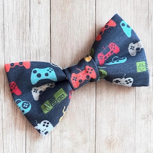 May include: A dark blue bow tie featuring a repeating pattern of colorful video game controllers in shades of red, light blue, white, and green. The fabric has a textured appearance and the design includes various handheld controller shapes.