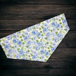 Puede incluir: Un camino de mesa de tela con un estampado floral de hortensias azules y hojas verdes sobre un fondo blanco. El camino de mesa tiene un borde cosido y está sobre un fondo de madera oscura. Las medidas son en centímetros.