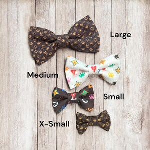 May include: Four bow ties in varying sizes labeled Large, Medium, Small, and X-Small. The Large and X-Small ties are dark brown with gold paw print patterns. The Medium tie is white with colorful paw prints and hearts. The Small tie is dark brown.