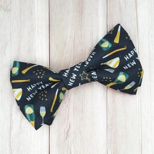 May include: A black bow tie featuring a repeating pattern of gold and white champagne flutes, party horns, and stars, with the text "HAPPY NEW YEAR" printed in white. The fabric has a textured finish and is shaped into a classic double-loop bow.
