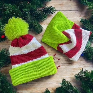 May include: A knitted beanie with a bright lime green pom-pom, featuring horizontal stripes of red, white, and red, and a wide lime green cuff. Beside it is a matching knitted neck warmer with a twisted design in lime green, white, and red.