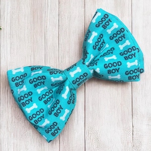 May include: A bright turquoise bow tie featuring a repeating pattern of white dog bones and the text GOOD BOY printed in a dark charcoal font. The fabric has a structured, folded shape with a central knot connecting the two sides of the bow tie.