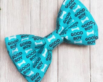 Puede incluir: Una pajarita de color turquesa brillante con un patrón repetitivo de huesos de perro blancos y el texto GOOD BOY impreso en una fuente gris oscuro. La tela tiene una forma estructurada y plegada con un nudo central que une ambos lados.