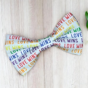 May include: A light blue bow tie featuring a repeating pattern of the words LOVE WINS printed in various colors including red, orange, yellow, green, blue, and purple. The fabric has a textured appearance and the bow tie is shaped in a classic style.