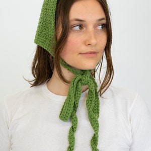 May include: A green crochet headscarf with long, twisted ties. The head covering is a solid, light green colour and is worn over the head and tied under the chin. The headscarf is made of a textured, crocheted material.