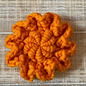 May include: A bright orange crocheted flower with a sparkly texture. The flower is made with a thick yarn and has a unique, textured design.