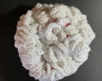 May include: A white crocheted scrunchie with silver metallic thread accents. The scrunchie is round and has a textured, ruffled appearance. The center reveals a hint of pink. This hair accessory is handmade.