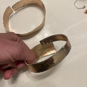 May include: Two gold-toned metal bracelets with a hammered finish. The bracelets are open and have a slight bend in them.