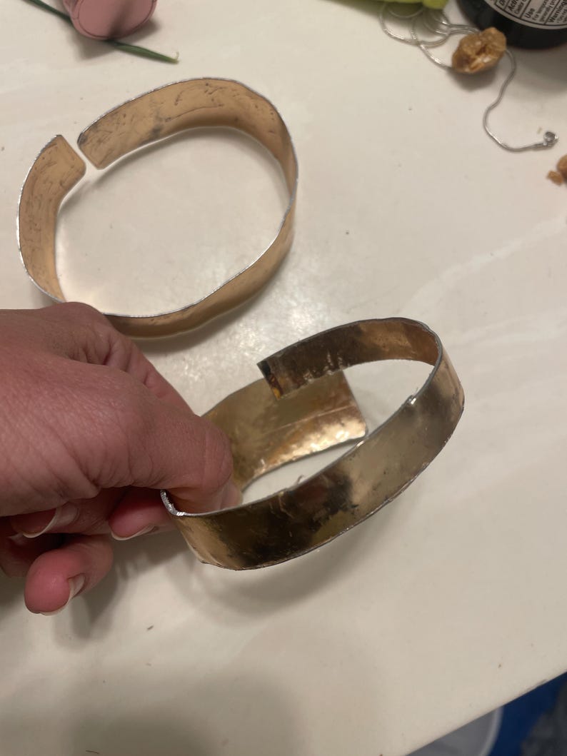 May include: Two gold-toned metal bracelets with a hammered finish. The bracelets are open and have a slight bend in them.