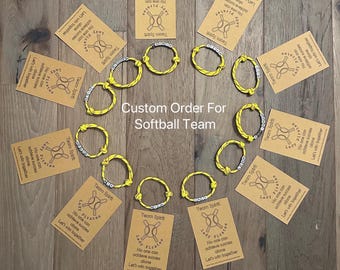 May include: A collection of yellow and silver bracelets arranged with accompanying cards on a wooden surface. The cards feature a softball-themed logo and text. The bracelets are custom-made for a softball team.