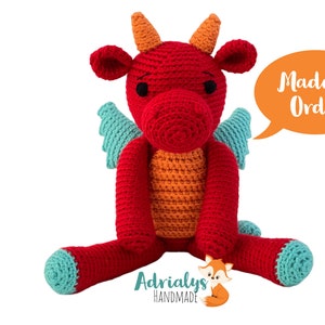 Puede incluir: Juguete de peluche de dragón rojo tejido a crochet con cuernos naranjas, alas turquesa y una burbuja de diálogo que dice "Hecho a pedido".
