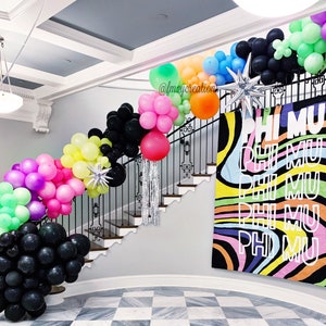 May include: A staircase decorated with colourful balloons, including black, green, pink, yellow, and blue. The balloons are arranged in a garland shape and are strung along the railing of the staircase. A large banner with the text "PHI MU" in white letters on a colourful background hangs on the wall behind the staircase.