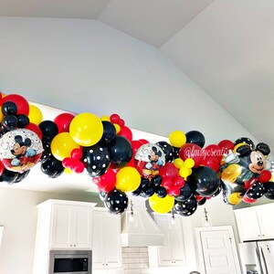 May include: A Mickey Mouse themed balloon garland in red, yellow, and black with Mickey Mouse character balloons. The garland is hanging from a ceiling in a room with white walls and cabinets.