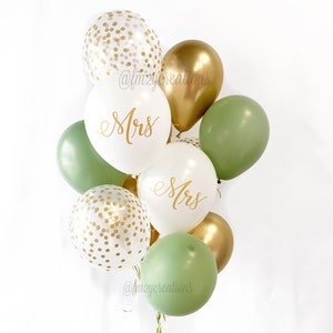 Puede incluir: Un ramo de globos verdes, blancos y dorados con la palabra "Mrs." escrita en dos de los globos blancos. Los globos están decorados con confeti dorado.