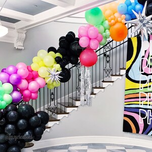 May include: A staircase decorated with colourful balloons in various shades of pink, green, yellow, and black. The balloons are arranged in a cascading pattern, creating a festive and celebratory atmosphere. The staircase is adorned with a black metal railing and a white wall. A colourful banner with the text "Peace, Peace, Peace" is displayed on the wall.