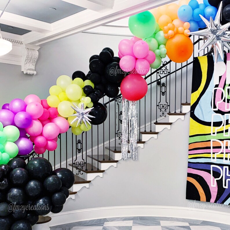 May include: A staircase decorated with colourful balloons in various shades of pink, green, yellow, and black. The balloons are arranged in a cascading pattern, creating a festive and celebratory atmosphere. The staircase is adorned with a black metal railing and a white wall. A colourful banner with the text "Peace, Peace, Peace" is displayed on the wall.