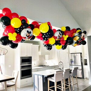 May include: A Mickey Mouse themed balloon garland in red, yellow, and black hangs from the ceiling of a kitchen. The garland is made of a variety of balloon sizes and shapes, including Mickey Mouse character balloons.