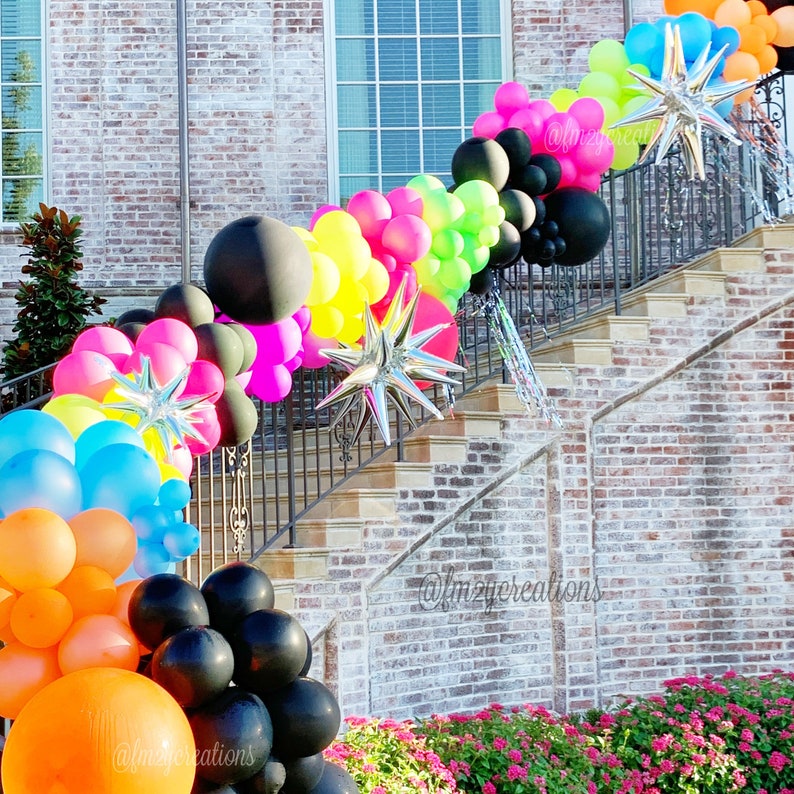 May include: A staircase decorated with a colourful balloon garland. The garland is made of black, pink, yellow, green, blue, and orange balloons. There are also silver star-shaped balloons and streamers. The staircase is made of brick and has a black metal railing.