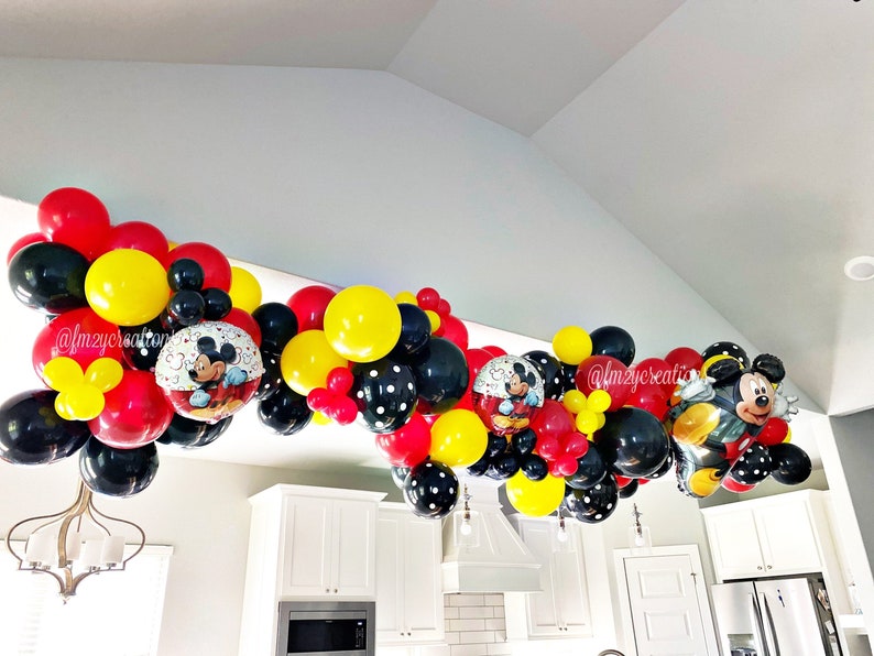 May include: A Mickey Mouse themed balloon garland in red, yellow, and black with Mickey Mouse character balloons. The garland is hanging from a ceiling in a room with white walls and cabinets.