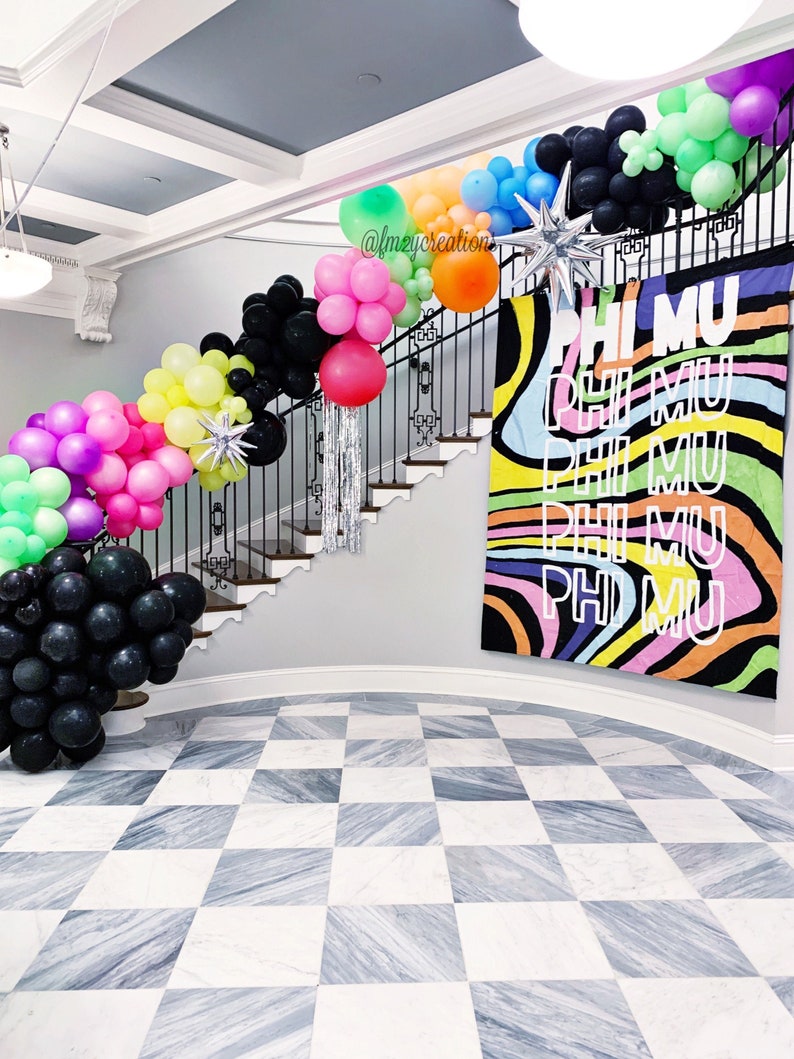 May include: A staircase decorated with colourful balloons, including black, green, pink, yellow, and blue. The balloons are arranged in a garland shape and are strung along the railing of the staircase. A large banner with the text "PHI MU" in white letters on a colourful background hangs on the wall behind the staircase.