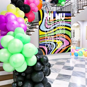 May include: A colourful balloon arch with black, green, pink, purple, yellow and blue balloons. The arch is in front of a staircase with a banner that says "PHI MU" in a rainbow swirl pattern.