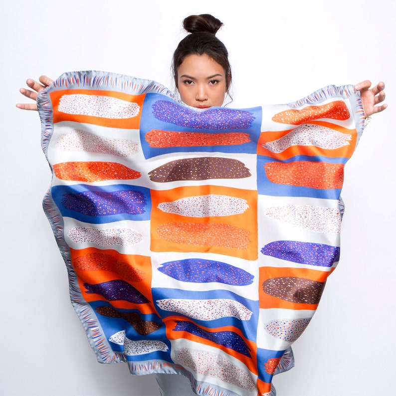 May include: A white, blue, orange and brown patterned silk scarf with a geometric design. The scarf is draped over a person's shoulders.