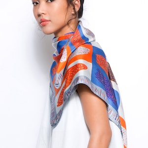 May include: A woman wearing a white top and blue jeans with a blue, orange and white patterned scarf tied around her neck.